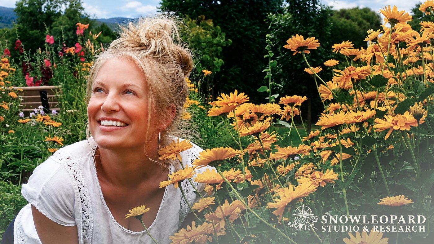 Kaitlyn Krahn smiling in a summer garden of golden flowers with mountains behind her, self portrait.