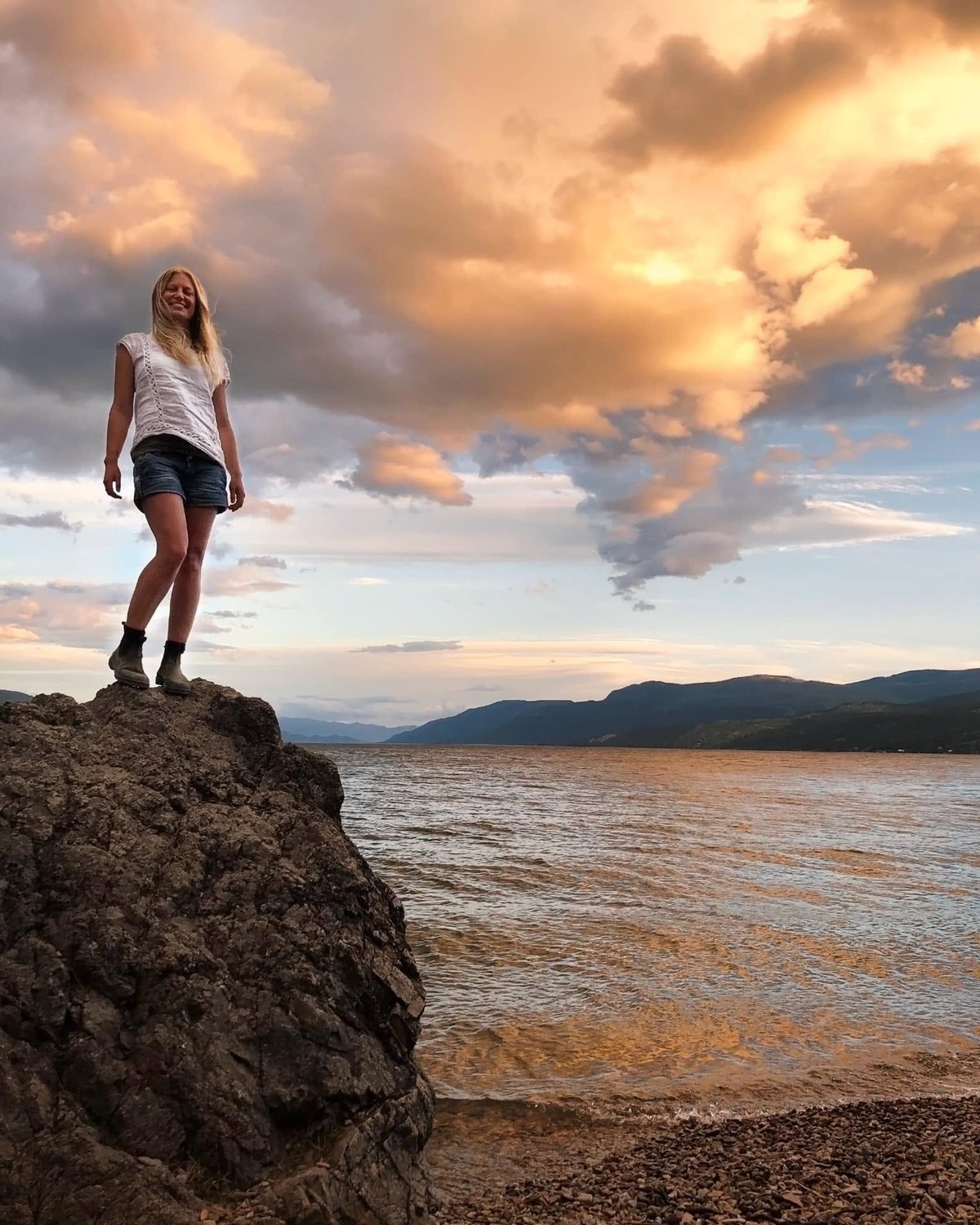 Kaitlyn Krahn standing on a rock above Okanagan Lake at sunset, the water and hills stretching behind her, self portrait.